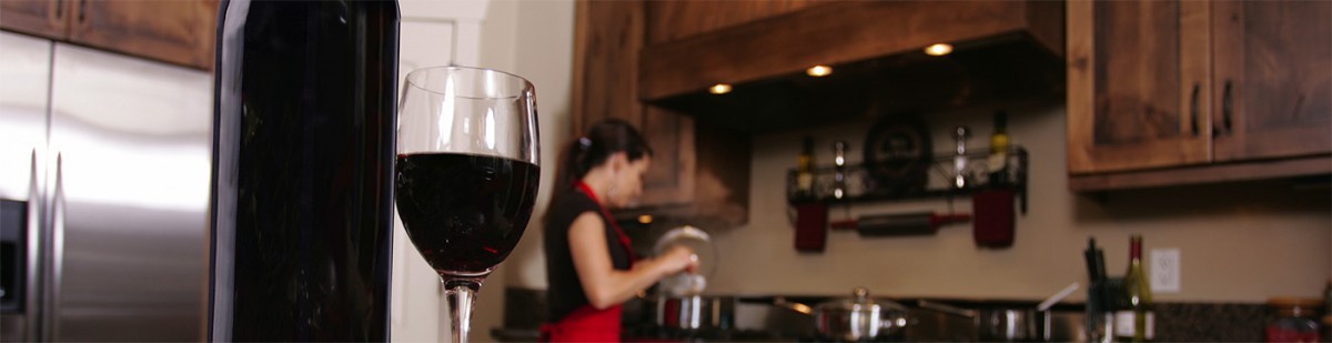 Glass of red wine and a bottle in the foreground of a kitchen where a woman in an apron cleans equipment