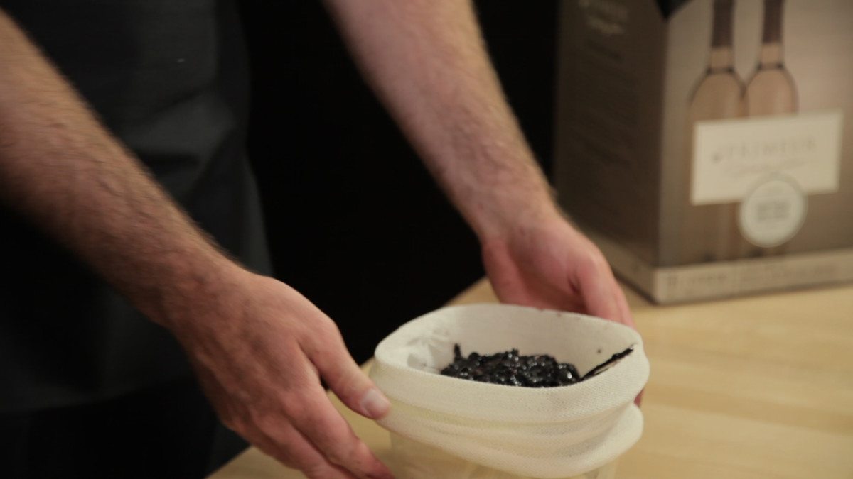 Des mains ouvrent un sac de trempage rempli de peaux de raisin foulées, devant une boîte de trousse En Primeur