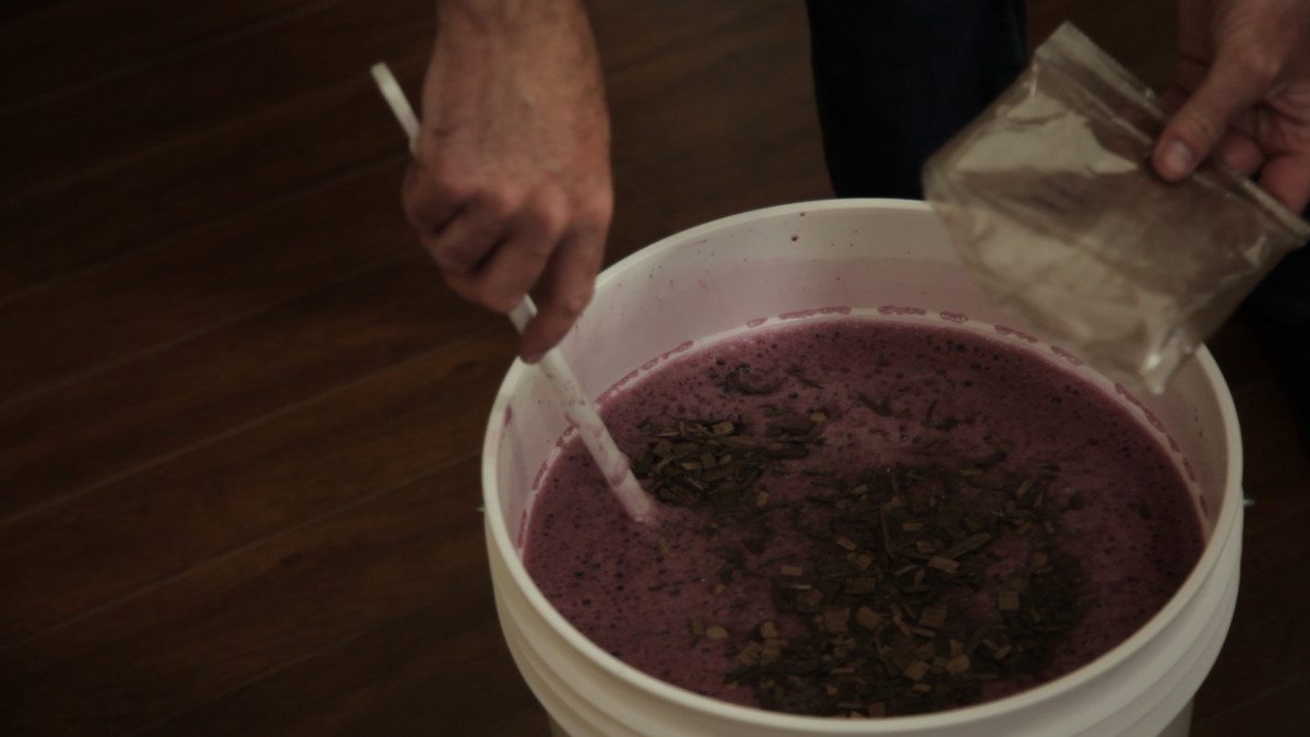 Des copeaux de chêne versés d'un sachet sur le moût violet d'une cuve de fermentation, remué à la spatule