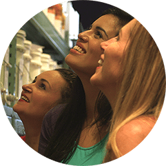 Three women smiling and looking up at shelves of winemaking supplies in a store