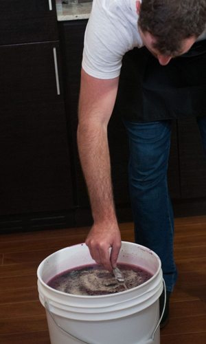 A man leaning over a white fermenting bucket, stirring frothy purple must with a spoon