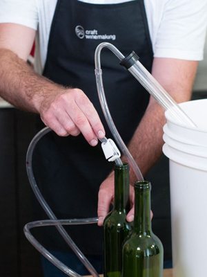 A winemaker holding a bottle-filling wand on a hose into a green wine bottle beside a stack of pails