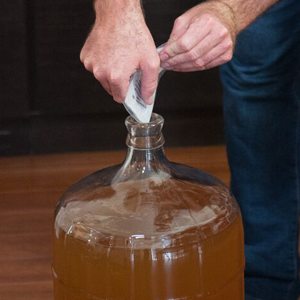 Hands tipping a sachet of clearing agent into the neck of a glass carboy of cloudy wine
