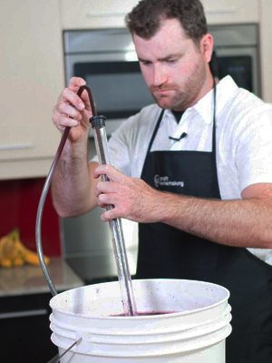 A winemaker lowering a racking cane and siphon hose into a bucket of red must