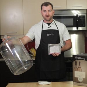 A winemaker in an RJS apron holding a glass carboy in one hand and a box of sanitiser in the other