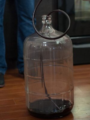 A drained glass carboy with a racking cane inside and a shallow layer of dark sediment left in the bottom