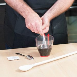 Hands stirring the contents of a sachet into a measuring jug of dark wine, with the empty sachet and a long spoon on the counter