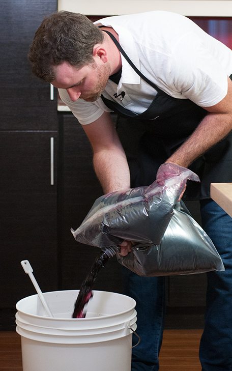 A winemaker in an apron pouring red grape juice from a foil bladder into a white fermenting bucket