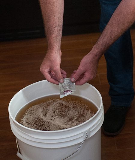 Hands emptying a sachet of dried yeast across the surface of juice in a white fermenting bucket