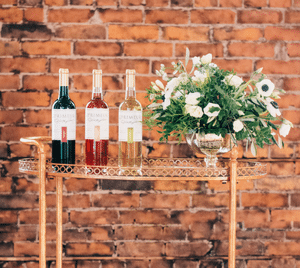 Three bottles of red, rose and white wine on a gold bar cart with white flowers, against a brick wall
