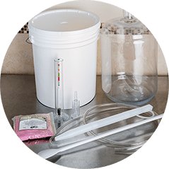 Winemaking equipment on a kitchen counter: a fermenting bucket with a hydrometer, a glass carboy, a siphon tube and a sanitiser sachet