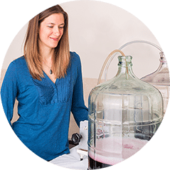 A smiling woman standing beside a large glass carboy fitted with a siphon tube
