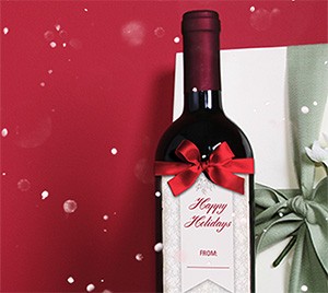 Bottle of red wine with a red bow and a Happy Holidays gift tag reading From, beside a wrapped present